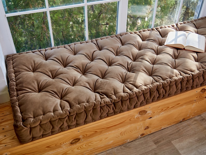 May include: A brown tufted cushion with a book on top, placed on a wooden window seat.