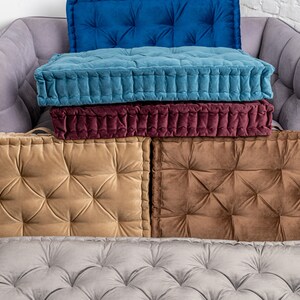 May include: Five floor cushions in different colours, each with tufted button detailing. The cushions are stacked on top of each other on a grey sofa.