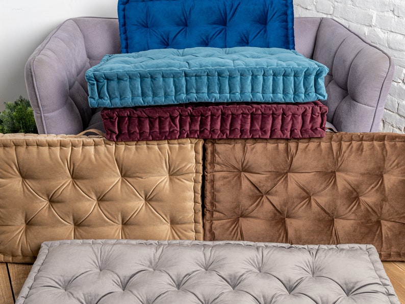 May include: Five floor cushions in different colours, each with tufted button detailing. The cushions are stacked on top of each other on a grey sofa.