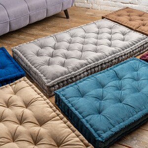 May include: Five floor cushions in different colours and textures. The cushions are rectangular and have a tufted design. The cushions are arranged in a row on a wooden floor.