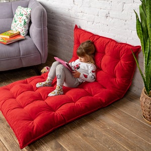 May include: A young girl sits on a red, tufted floor cushion and uses a pink tablet. The cushion is on a wooden floor. A gray sofa and a potted plant are in the background.
