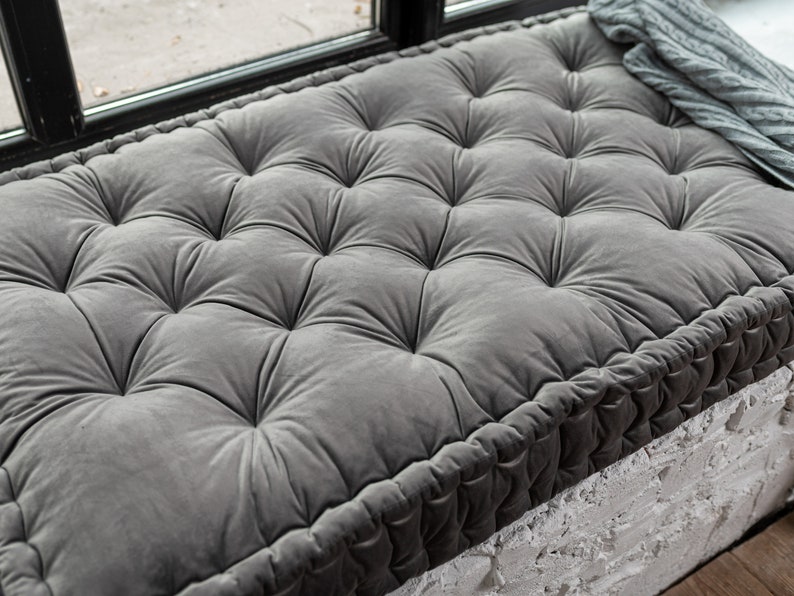 May include: A grey tufted floor cushion with a thick, rope-like border. The cushion is made of a soft, plush fabric and is perfect for adding comfort to any room.