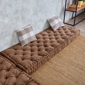 May include: Three brown floor cushions with tufted buttons, arranged in a row against a concrete wall. Each cushion has a white and grey checkered pillow on top.