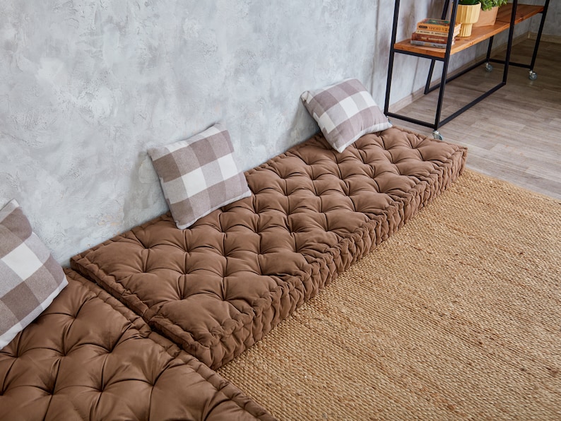 May include: Three brown floor cushions with tufted buttons, arranged in a row against a concrete wall. Each cushion has a white and grey checkered pillow on top.