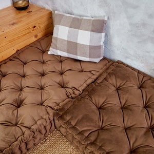 May include: Two brown tufted floor cushions with a white and brown checkered pillow on top. The cushions are placed on a wooden platform with a light brown wood grain.
