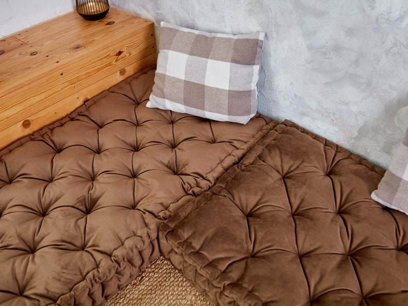 May include: Two brown tufted floor cushions with a white and brown checkered pillow on top. The cushions are placed on a wooden platform with a light brown wood grain.