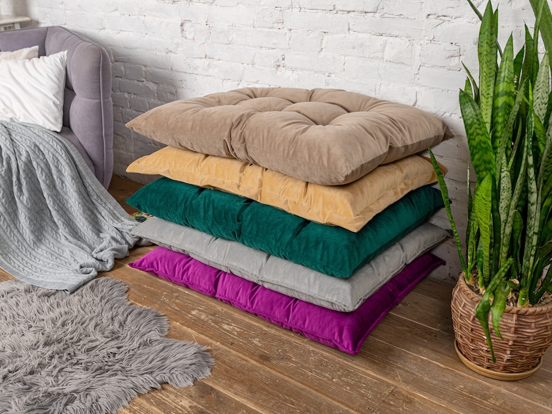May include: A stack of five cushions in various colors, including beige, yellow, green, gray, and purple. The cushions are arranged in a pyramid shape on a wooden floor.