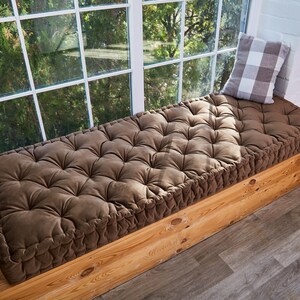 May include: A brown tufted window seat cushion with a white and grey checked pillow on top. The cushion is placed on a wooden window seat.