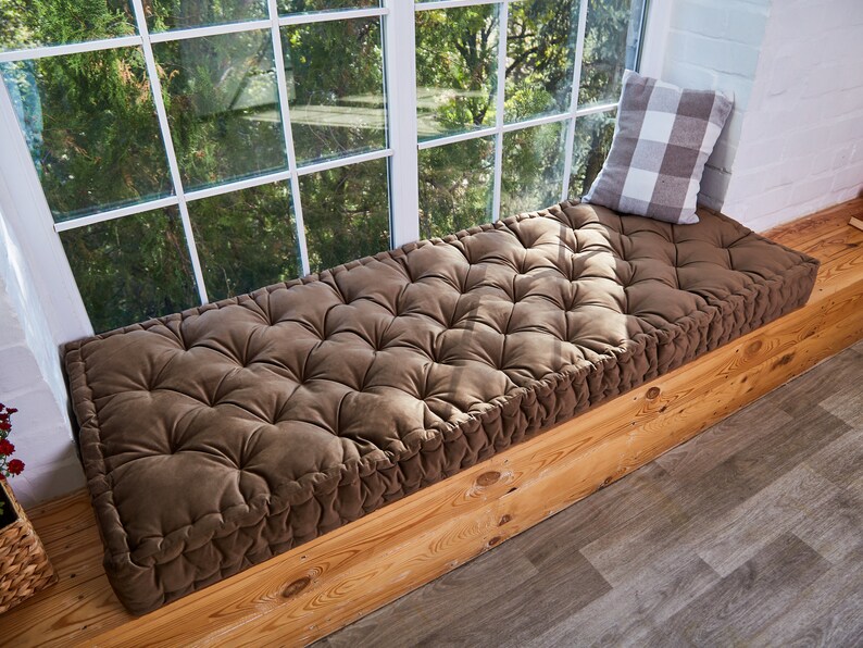 May include: A brown tufted window seat cushion with a white and grey checked pillow on top. The cushion is placed on a wooden window seat.