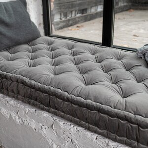May include: A grey tufted window seat cushion with a grey throw blanket on top. The cushion is made of a soft, plush fabric and has a thick, comfortable padding.