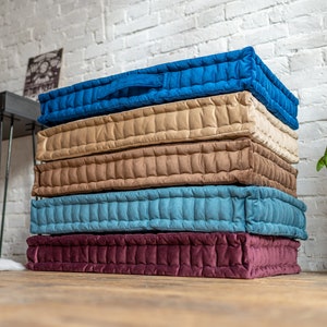 May include: A stack of five floor cushions in different colours. The cushions are made of a quilted fabric and have a tufted design. The colours are burgundy, blue, beige, brown, and blue.