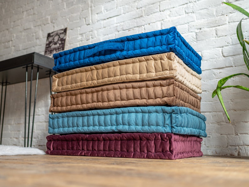 May include: A stack of five floor cushions in different colours. The cushions are made of a quilted fabric and have a tufted design. The colours are burgundy, blue, beige, brown, and blue.