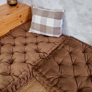 May include: Two brown tufted floor cushions with a white and brown checked pillow on top. The cushions are placed on a wooden platform with a light brown wood grain.