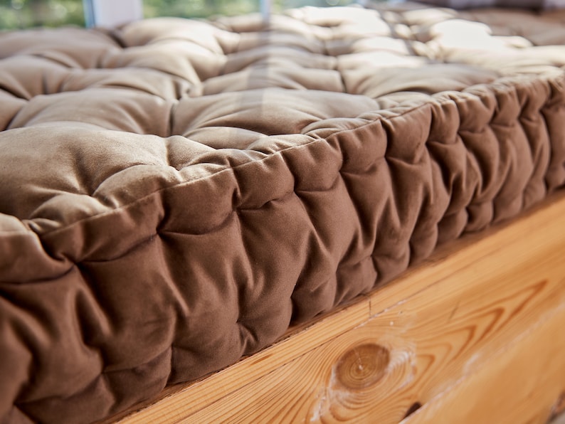 May include: Close-up of a brown tufted cushion with a thick, soft fabric. The cushion is placed on a wooden surface.