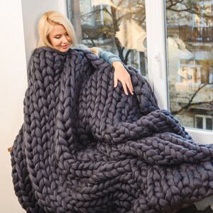 Può includere: Una donna seduta vicino a una finestra avvolta in una grande coperta di maglia grigia spessa. La coperta è realizzata con filato spesso e presenta un motivo a treccia.