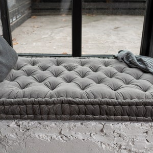 May include: A grey tufted cushion with a thick, grey fabric border sits on a window seat. The cushion is made of a soft, plush material and has a comfortable, inviting look.