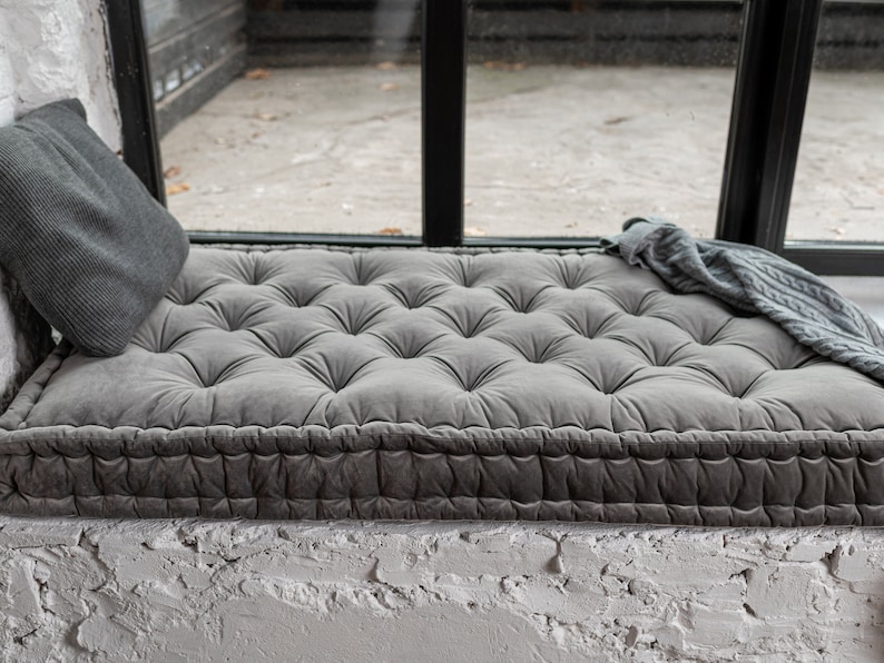 May include: A grey tufted cushion with a thick, grey fabric border sits on a window seat. The cushion is made of a soft, plush material and has a comfortable, inviting look.
