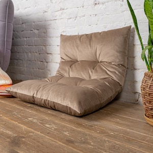 May include: A brown velvet floor cushion with a tufted design. The cushion is sitting on a wooden floor in front of a white brick wall.