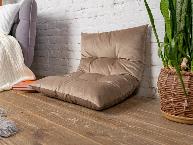 May include: A brown velvet floor cushion with a tufted design. The cushion is sitting on a wooden floor in front of a white brick wall.