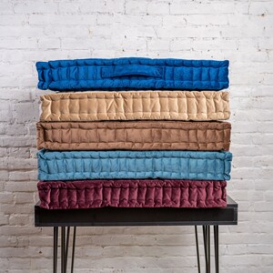 May include: A stack of five quilted floor cushions in various colours, including beige, brown, blue, and burgundy. The cushions are arranged on a black metal table with hairpin legs.