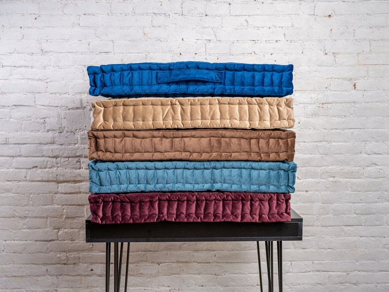 May include: A stack of five quilted floor cushions in various colours, including beige, brown, blue, and burgundy. The cushions are arranged on a black metal table with hairpin legs.