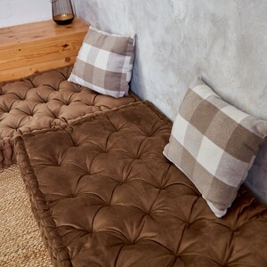 May include: Two brown floor cushions with button tufting, one on top of the other, with a white and brown checked pillow on each cushion.