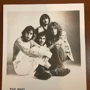 May include: Black and white promotional photo of the band The Who. The band members are all wearing casual clothing and are looking at the camera. The photo is captioned "THE WHO" and ".MCA RECORDS".
