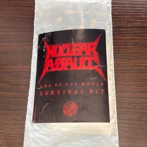 以下が含まれることがあります： 白のプラスチック製の袋に、黒と赤のステッカーが貼られています。ステッカーには、太字の赤いフォントで「Nuclear Assault」というテキストが書かれています。テキストの下には、白いテキストで「End of the World Survival Kit」と書かれています。赤い円の中に、白いテキストで「In-Effect」と書かれています。