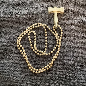 Puede incluir: Un collar con colgante dorado con un colgante en forma de martillo. El colgante está unido a una cadena de bolas. El collar se muestra sobre una superficie texturizada de color gris oscuro.