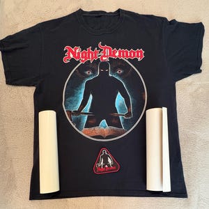 May include: Black t-shirt with a graphic featuring the text "Night Demon" in red, a silhouetted figure, and a wolf. Includes a patch with the same logo and two rolled-up items.