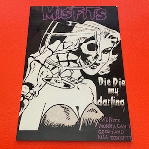 May include: Black and white graphic art print featuring the word "MISFITS" in purple. The image depicts a skeleton holding a wine glass with a woman's face. Text includes "Die Die my darling" and "We bite mommy, can I go out and kill tonight?"