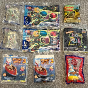 May include: A collection of ten colorful plastic bags featuring various cartoon characters. The bags include characters from 'Curious George', 'Chalk Zone', 'Stuart Little', and 'Mr. Potato Head'.