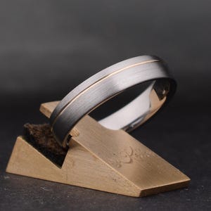 May include: A silver ring with a gold inner band sits on a geometric display stand. The ring features a brushed metal finish and a thin gold stripe. The stand is made of gold-coloured metal and has a dark, textured base.