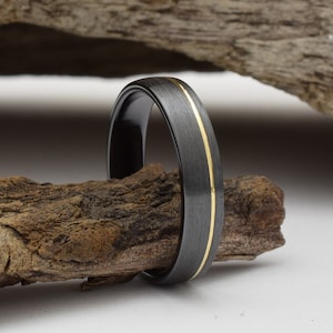 Op de afbeelding: Een zwarte en gouden trouwring met een geborstelde afwerking. De ring heeft een dunne gouden lijn rond de rand.