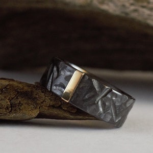 May include: A black and gold ring with a textured band and a thin gold inlay. The ring is resting on a piece of driftwood.