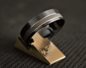 May include: A black and silver wedding band with a brushed finish. The band is on a gold display stand.