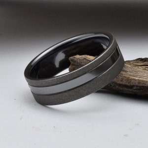 May include: A black and gray wedding band with a brushed finish and a polished black inlay. The band is made of tungsten carbide and is a popular choice for men's wedding rings.