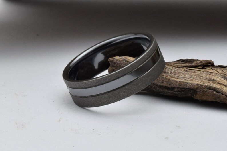 May include: A black and gray wedding band with a brushed finish and a polished black inlay. The band is made of tungsten carbide and is a popular choice for men's wedding rings.