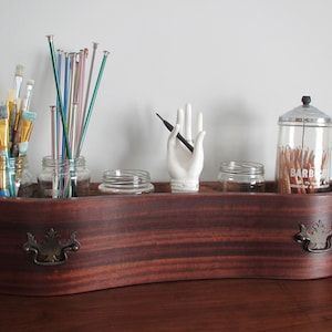 Könnte beinhalten: Eine Holzkiste mit einer gewölbten Vorderseite, die mit zwei Metallgriffen ausgestattet ist. Die Kiste ist mit verschiedenen Gläsern gefüllt, die Kunstzubehör wie Pinsel, Stricknadeln und Bleistifte enthalten. Eine weiße Keramik-Handfigur hält einen schwarzen Bleistift.