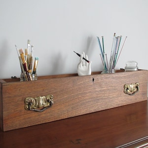 Könnte beinhalten: Eine Holzkiste, die als Aufbewahrungsorganisator mit zwei Messinggriffen umfunktioniert wurde. Die Kiste ist gefüllt mit Gläsern mit Pinseln, Stricknadeln und einer Keramikhand, die einen Bleistift hält.