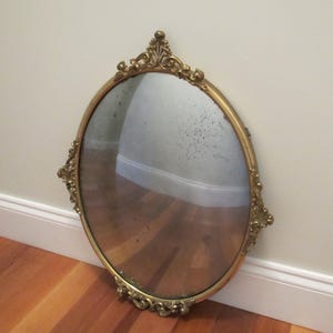 Puede incluir: Espejo antiguo ovalado con marco dorado ornamentado. El espejo está apoyado contra una pared blanca y un suelo de madera marrón. El marco presenta adornos decorativos en la parte superior, inferior y laterales. La superficie del espejo muestra signos de edad.