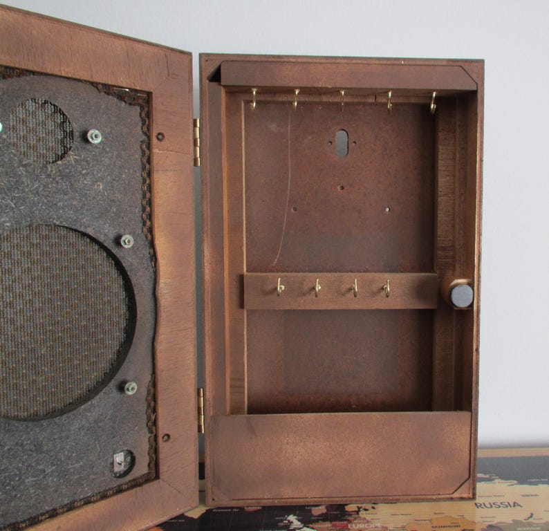 May include: A vintage wooden key cabinet with a dark brown finish. The cabinet has a hinged door with a speaker-like design on the left side and key hooks inside. The cabinet is open, revealing the interior with key hooks and a magnetic closure.