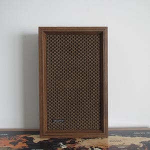 May include: A vintage Sony speaker with a brown wooden frame and a dark brown mesh speaker cover. The speaker has a rectangular shape and is designed for audio playback. The Sony logo is visible on the speaker.