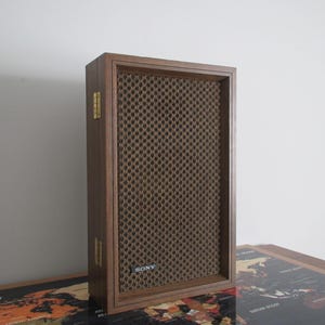May include: A vintage Sony speaker with a dark wood frame and a brown mesh front. The speaker has a rectangular shape and gold-colored hinges. The Sony logo is visible on the lower front of the speaker.