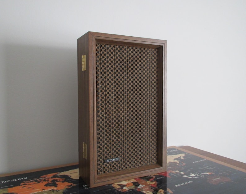 May include: A vintage Sony speaker with a dark wood frame and a brown mesh front. The speaker has a rectangular shape and gold-colored hinges. The Sony logo is visible on the lower front of the speaker.