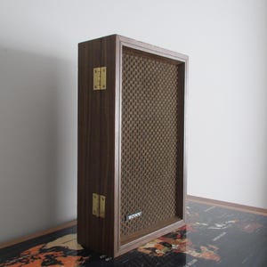 May include: A vintage Sony speaker with a dark wood frame and a brown mesh front. The speaker has brass-colored hinges and the Sony logo is visible on the lower right. The speaker is rectangular and stands upright.