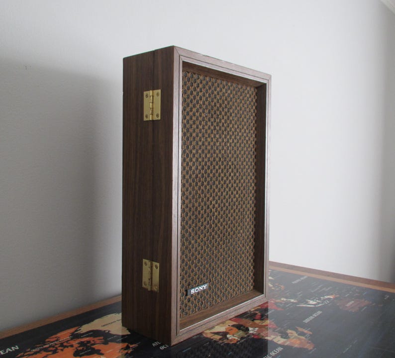May include: A vintage Sony speaker with a dark wood frame and a brown mesh front. The speaker has brass-colored hinges and the Sony logo is visible on the lower right. The speaker is rectangular and stands upright.