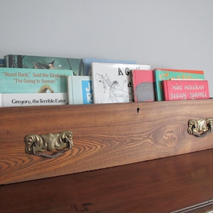 Könnte beinhalten: Eine Holzkiste, die als Bücherregal umfunktioniert wurde, mit einem Messinggriff und mehreren Kinderbüchern, die darin gestapelt sind. Die Bücher tragen Titel wie "Stand Back, Said the Elephant, I'm Going to Sneeze!" und "Gregory, the Terrible Eater".