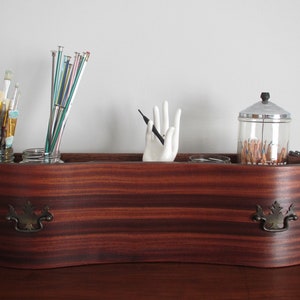 Könnte beinhalten: Ein Holzorganizer mit drei Fächern, die jeweils ein Glasgefäß mit Bastelbedarf enthalten. Der Organizer hat zwei Messinggriffe und eine gewellte Oberkante. Die Gläser enthalten Pinsel, Stricknadeln und Bleistifte.