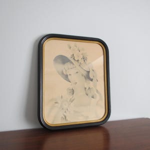 May include: Framed vintage print of a woman wearing a hat adorned with flowers and ribbons. The artwork is in a black and gold frame, resting on a dark wooden surface. The print has a sepia tone, giving it a classic, antique aesthetic.